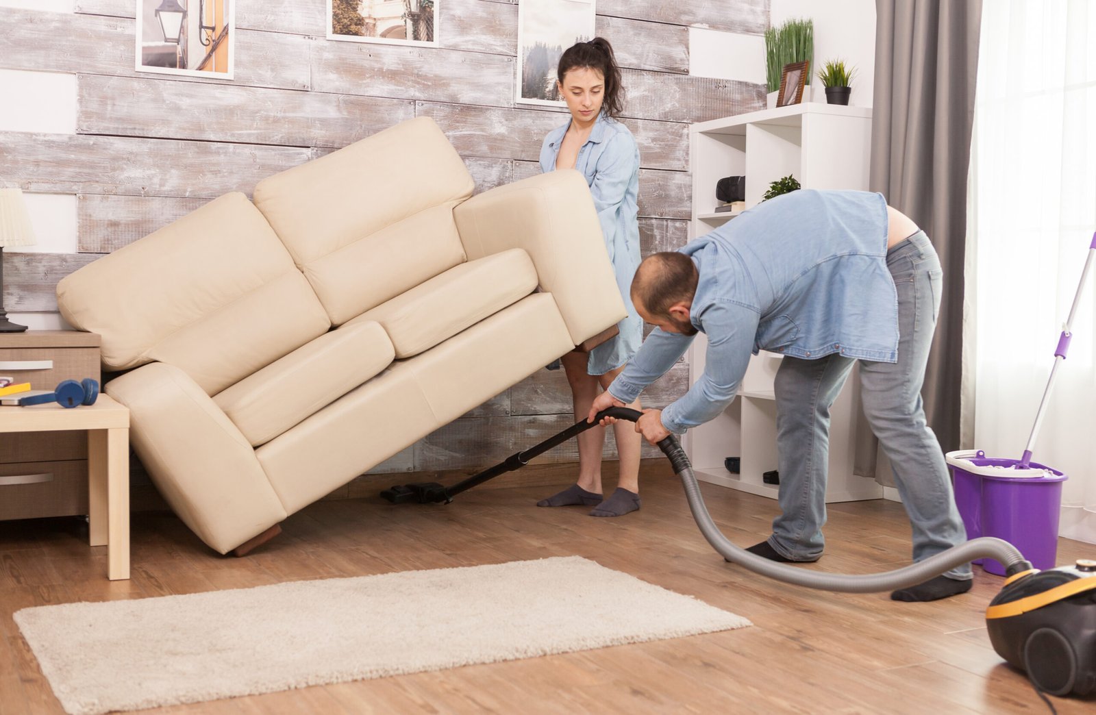 Wife picks up sofa while her husband is cleaning the dust under it with vacuum cleaner.